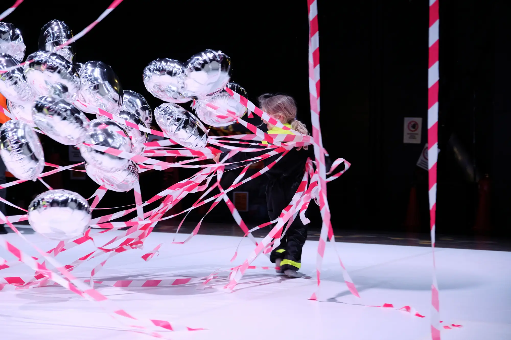 The photo shows a child playing and dancing with silver helium balloons, with red-and-white ribbons dangling from them.
