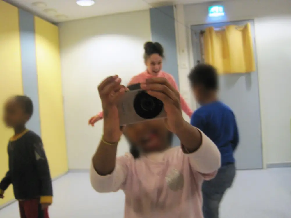 A photograph showing a child taking a picture, with a researcher and another child dancing in the background.