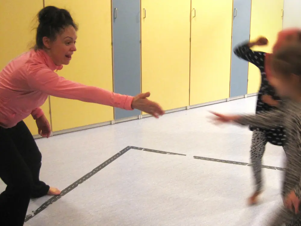 A photograph showing a researcher and a child reaching out toward each other in the multipurpose room of a daycare center. Blue and yellow cabinets are visible in the background.