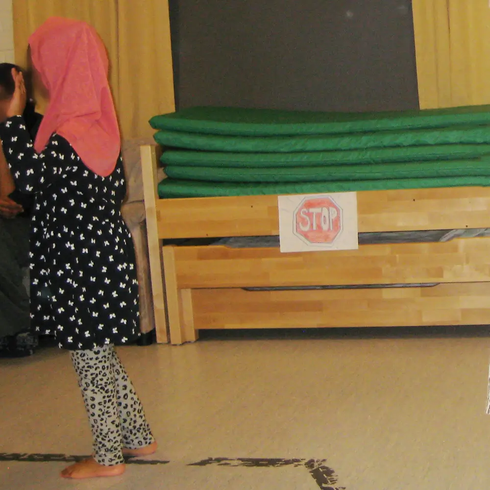 The photograph shows a child standing barefoot indoors on a light-colored floor. In the background are stacked green mattresses on a wooden bench with a hand-drawn red STOP sign.