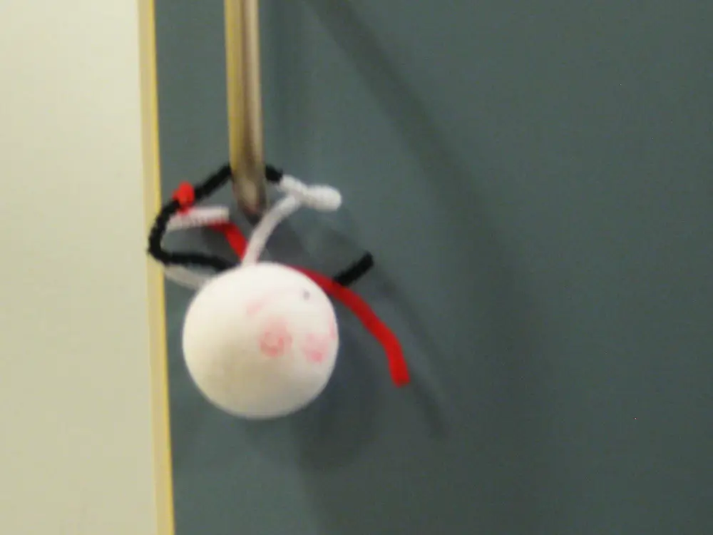 A photograph of a white paper ball with facial features drawn on it in pink marker. Black, red, and white pipe cleaners are attached to the ball as legs, and the ball is hung from a doorknob.