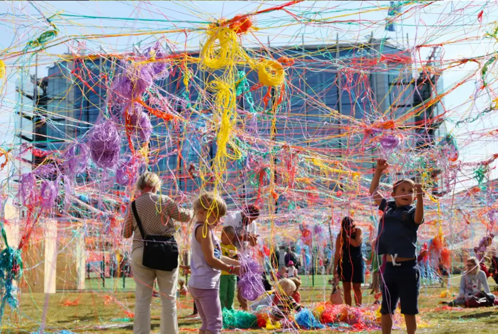 The photograph shows an outdoor artwork made of colourful threads, through which children and adults walk and touch. The piece consists of red, yellow, purple, and orange threads that form a complex and tangled network. Photo by Karoline H Larsen 2014.