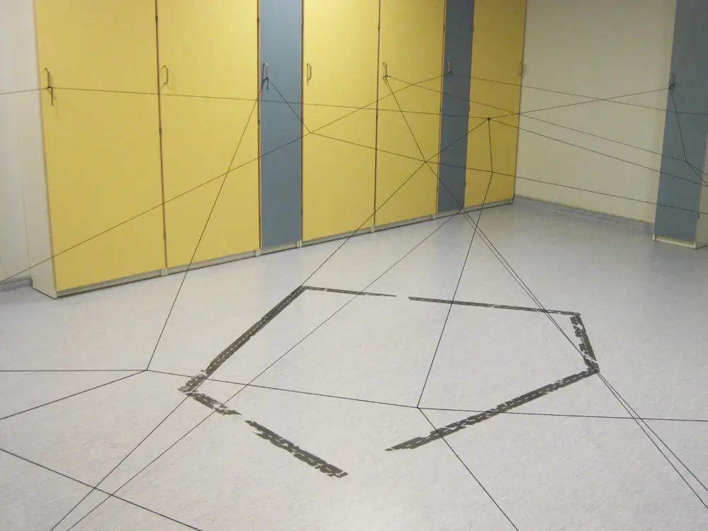 The photograph shows an empty multipurpose room in a daycare centre, featuring yellow and blue cabinets and an irregular polygon outlined on the floor with black tape. The room contains a net made of thin black threads that extends from the cabinets to the floor, creating a three-dimensional structure.