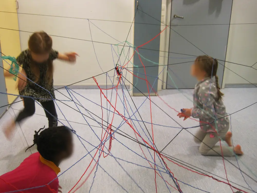 The photograph shows three children playing in a space filled with a complex web of multicoloured threads. Red, blue, and green threads form a lattice-like structure beneath and through which the children move.