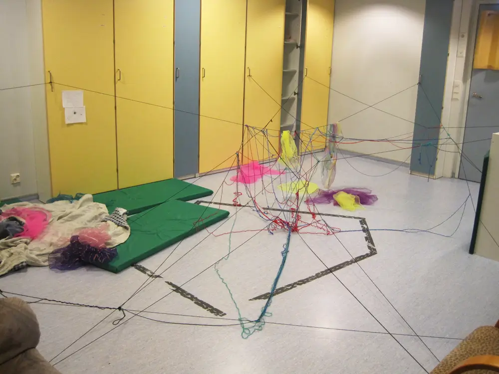 A photo of a messy multipurpose room at a daycare centre, with multicoloured strings forming a partly broken web on the floor and between the furniture. The room features yellow and blue cabinets, green mattresses, and colorful chiffon scarves.