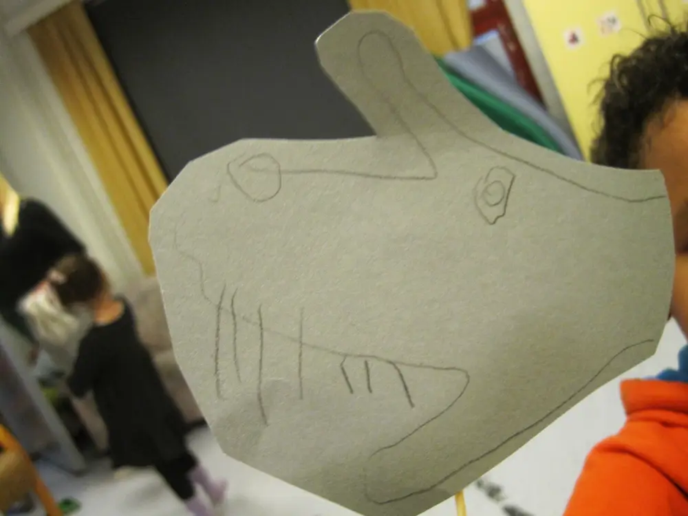 A photograph showing a child presenting a character they have drawn that resembles an animal&rsquo;s head. The artwork was created by tracing the outline of the child&rsquo;s palm onto paper and adding an eye, a nose, and teeth to the drawing.
