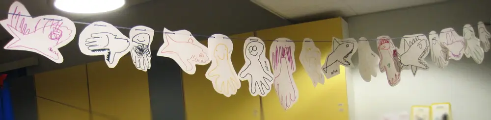 The photo shows the daycare centre&rsquo;s multipurpose room, with a string stretched high across the room and several animal figures of different shapes and colours strung along it. The figures are created by tracing a child&rsquo;s hand onto paper, adding eyes, mouths, teeth, and other features, and then cutting them out.