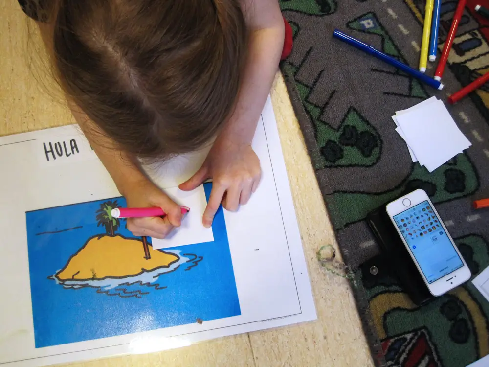 A photo of a child drawing on a small piece of paper on a drawing pad on the floor. The photo also shows a smartphone with emojis on the screen and felt-tip pens on the floor.
