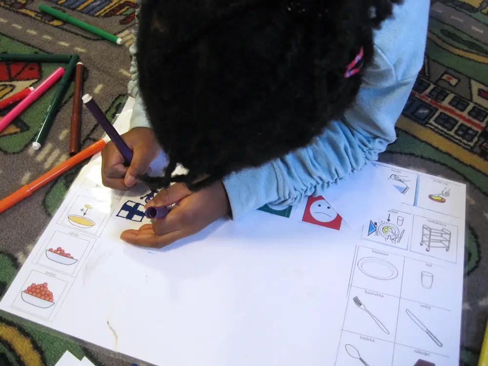 A photo of a child drawing a Finnish flag and people on a small piece of paper on a drawing pad on the floor. The photo also shows felt-tip pens on the floor.