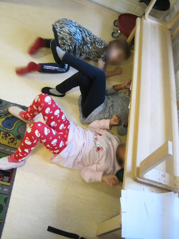 The photograph shows three children on the floor who have crawled under a long wooden bench in the daycare centre&rsquo;s multipurpose room.