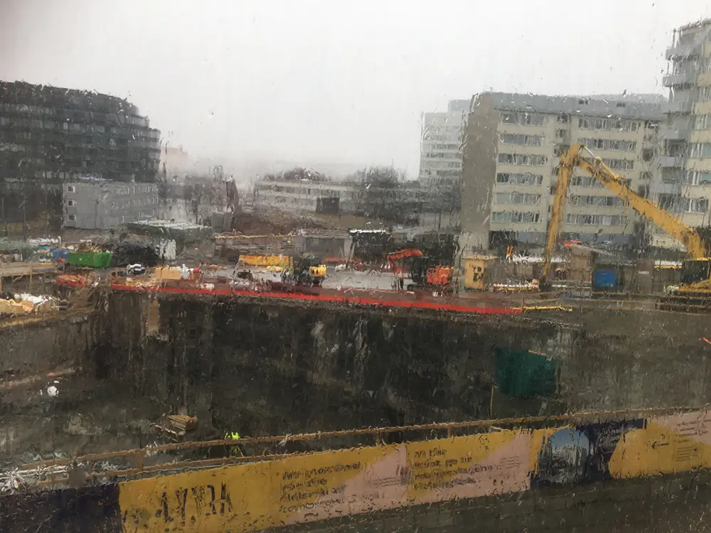 A construction site in an urban area on a rainy day, showing an excavation pit, construction equipment, and apartment buildings in the background. Through the rain-streaked window, the photo shows the construction site fence, a crane, and other construction materials.