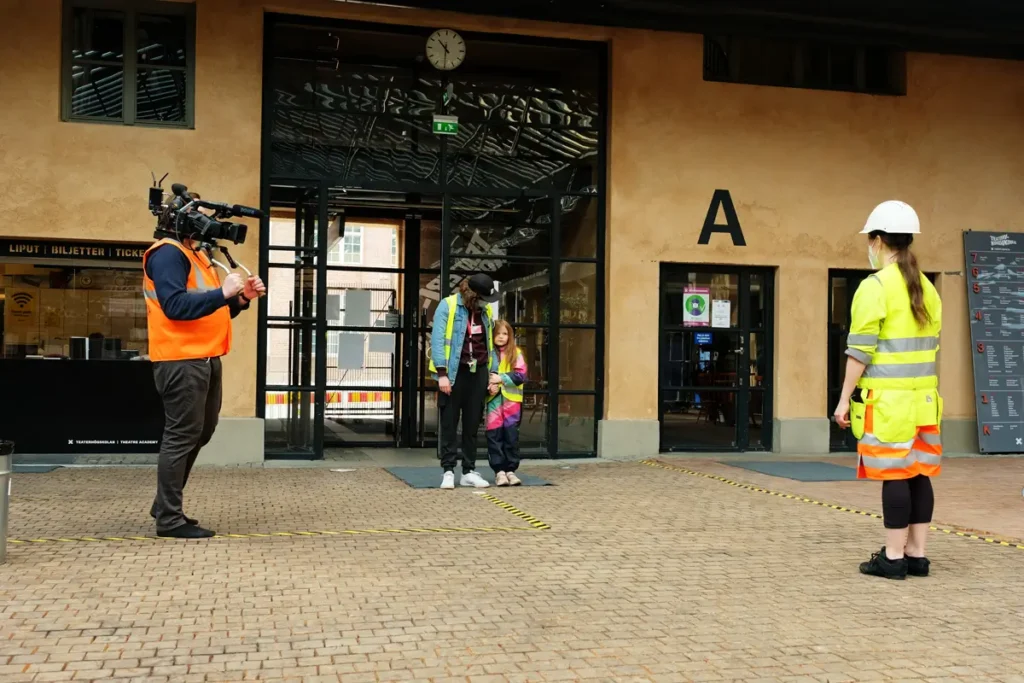 The photo shows the area in front of the entrance to the Theatre Academy, where a person with a camera is photographing a researcher in construction gear, as well as a daycare teacher and a preschool-aged child. The individuals are maintaining the recommended social distance of more than two meters during the COVID-19 pandemic.