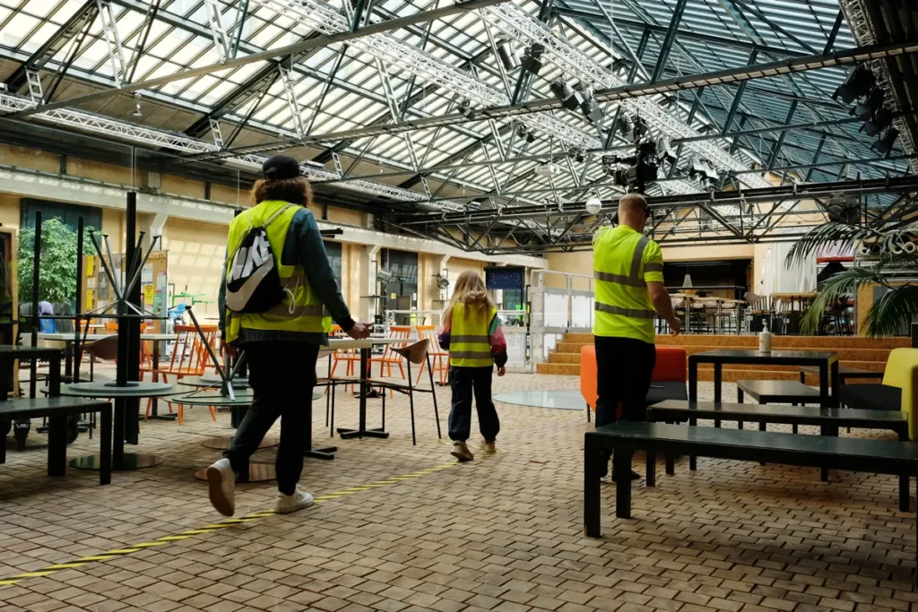 A photograph of a child and an adult walking one behind the other along a path marked with yellow-and-black safety tape on the floor of the Theatre Academy&rsquo;s lobby, which features a high glass ceiling and metal structures. The subjects are wearing yellow reflective vests, and the space contains tables and chairs.