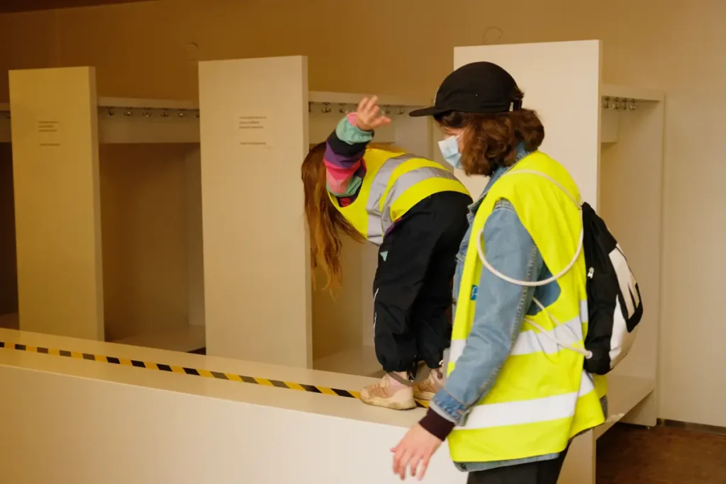 A photograph of a child and an adult wearing high-visibility vests and face masks indoors. The child is standing on a raised platform, bent over along a path marked with tape; the adult is standing beside them, with empty coat hooks visible in the background.