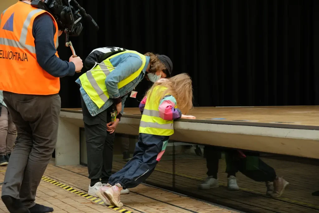 The photograph shows a child and an adult examining photographs attached to the floor of the stage at the Theatre Academy lobby.