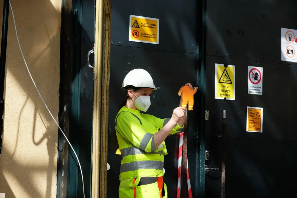 A photograph taken outside the studio, with preparations for the event underway, showing three people wearing yellow and white safety helmets and reflective vests. The researcher stands in front of the doorway holding a drawn cartoon-style orange jumpsuit. Warning signs and a closed black door are visible in the background.