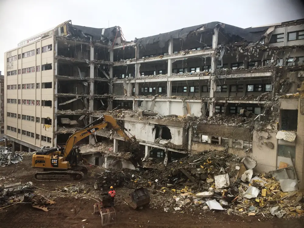 The photograph shows a partially demolished office building, revealing its interior and collapsed structures. The image shows a yellow excavator demolishing the building, surrounded by a pile of construction debris and rubble.