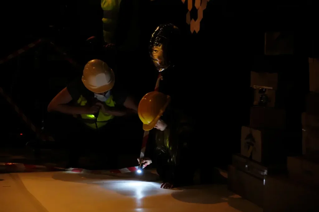 A photo collage showing, on the left, two construction helmets and two flashlights on the floor of the event space, with photographs and red string attached to them. On the right, a child and an adult, both wearing helmets, are examining the drawings on the floor by flashlight in the dark.