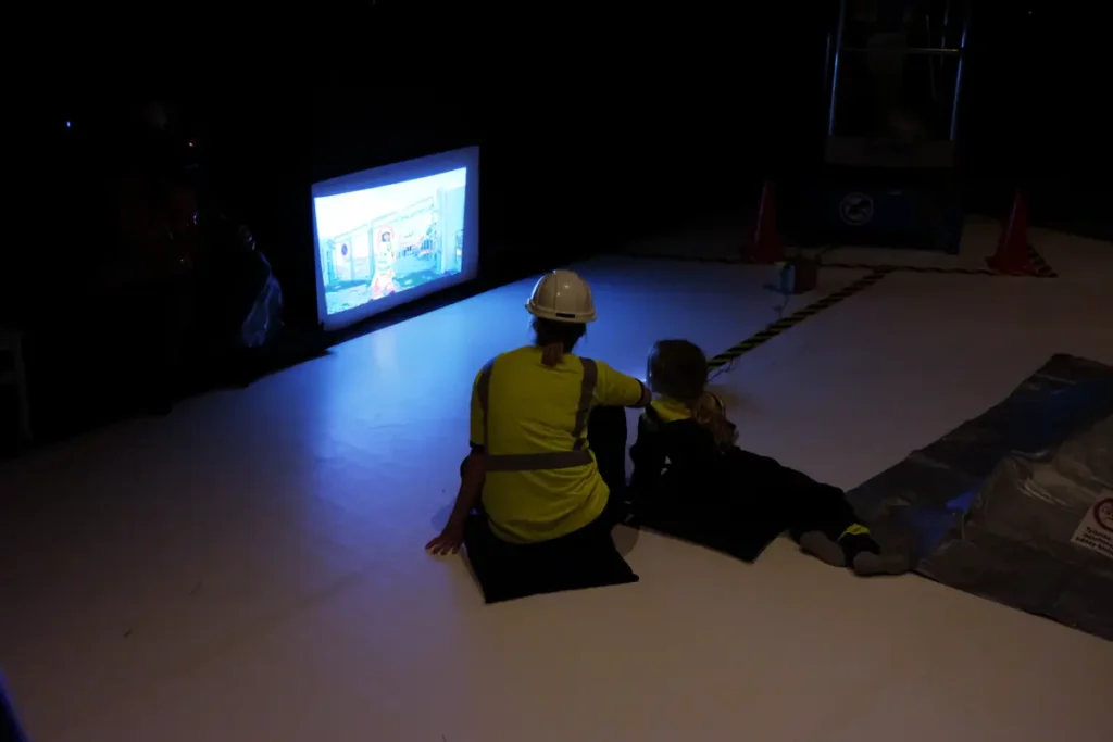 A collage of four photographs depicting the video-viewing setup. The first two images show the video projected from a small projector attached to a blue Duplo truck. The screen is mounted on the wall at floor level, with the words &ldquo;Muutostilassa &ndash; In Transformation&rdquo; projected onto it. The other two photographs show a child and an adult seated on large floor cushions, watching the screen in a dark room.