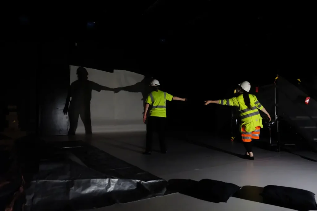 This collage of two photographs depicts a play of shadows in a dimly lit space, where the shadows of two people are cast onto a screen on the wall.
