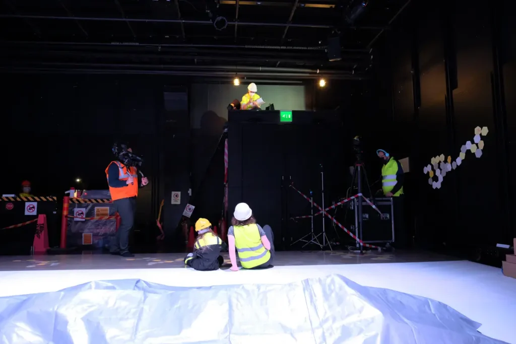 The photograph depicts a scene in which the researcher is sitting on a loft built above the doorway, while the participating child and adult are seated on the floor in front of it. In addition, one person is filming the scene with a video camera, and another is handling the camera equipment. The room features black walls, technical equipment, as well as caution tape and warning signs.