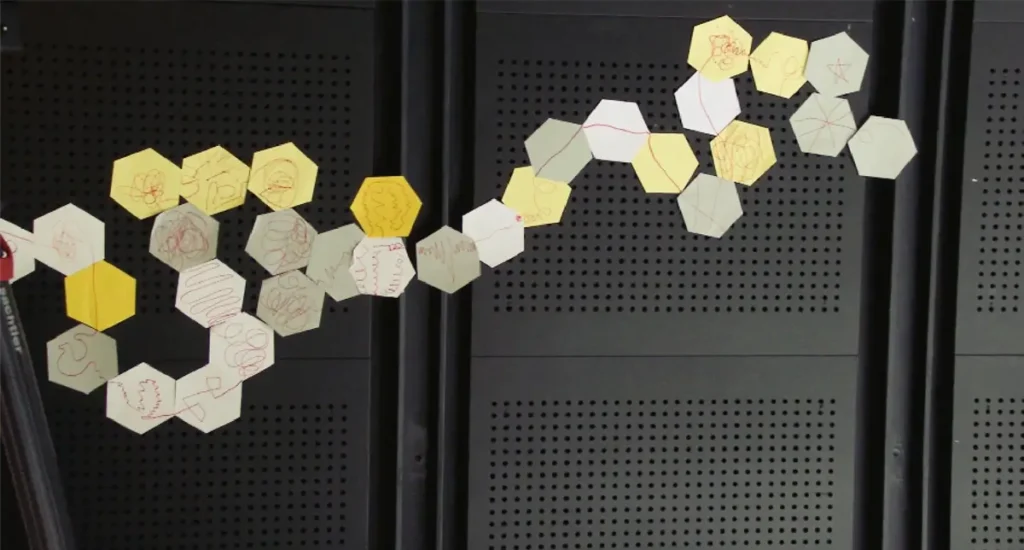 A photograph showing 31 white, yellow, and grey hexagonal pieces of paper connected to one another. Various lines have been drawn on the hexagons, and the hexagons are connected to one another in such a way that the lines from different hexagons intersect.