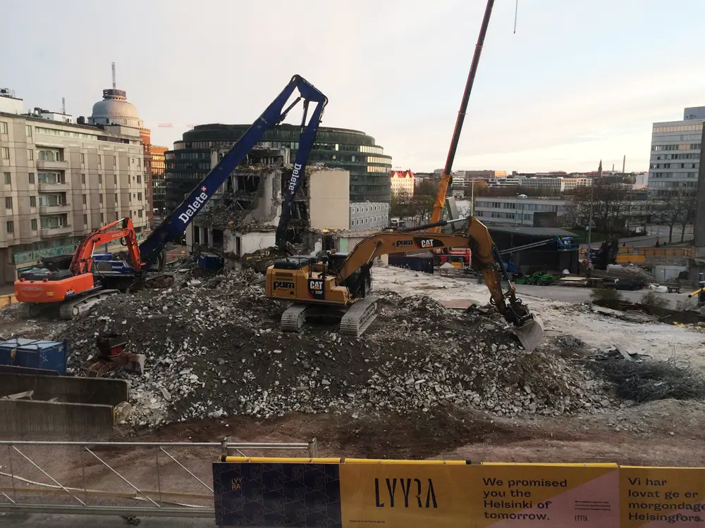 A construction site where two excavators are demolishing an old building and piling up rubble and concrete debris. A cityscape is visible in the background, and in the foreground is a construction site fence bearing the advertising text &ldquo;LYYRA&rdquo;.