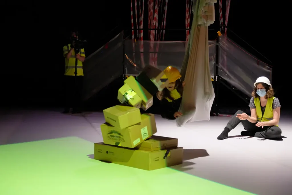 The photo shows a child knocking over a tower of cardboard boxes. The child is inside an aerial yoga hammock at floor level. An adult is seated nearby and has given the child a push, causing the child to hit the cardboard box tower and knock it over. In the background, a third person wearing a yellow safety vest is recording the scene with a video camera.