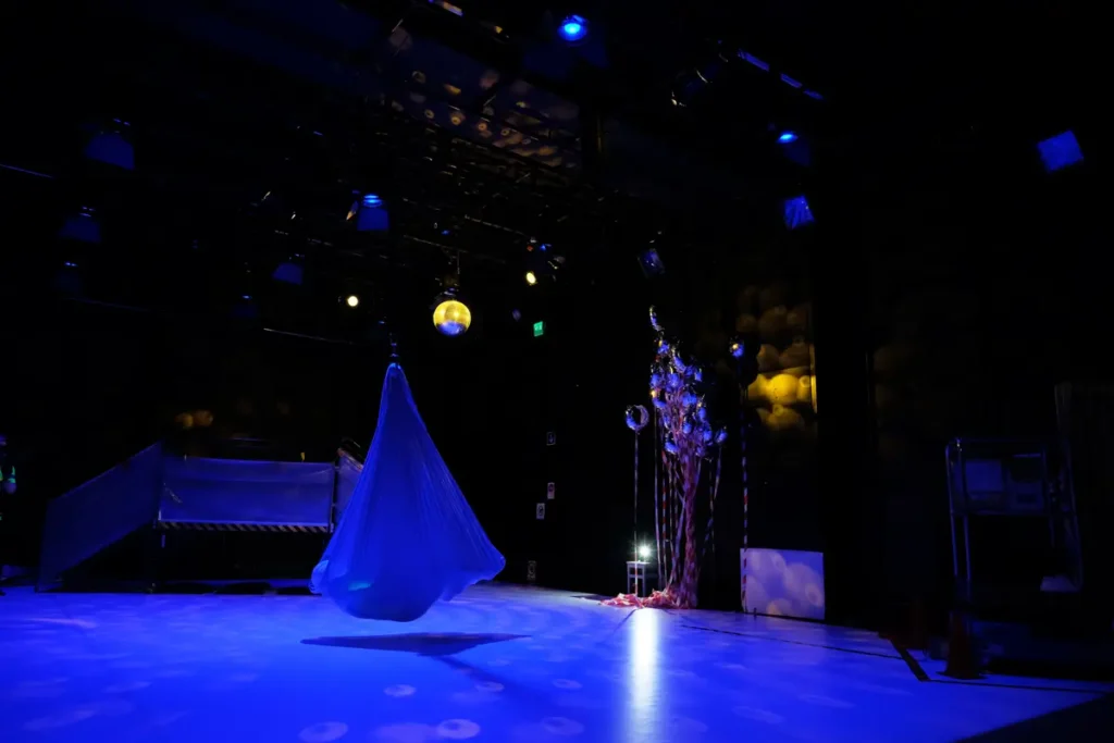 The pair of photos depicts a serene scene lit by dark blue theatre lighting, with an aerial yoga hammock at the centre of the room. In the photo on the left, a child lies inside the hammock while an adult sits nearby watching. In the photo on the right, both are inside the hammock.