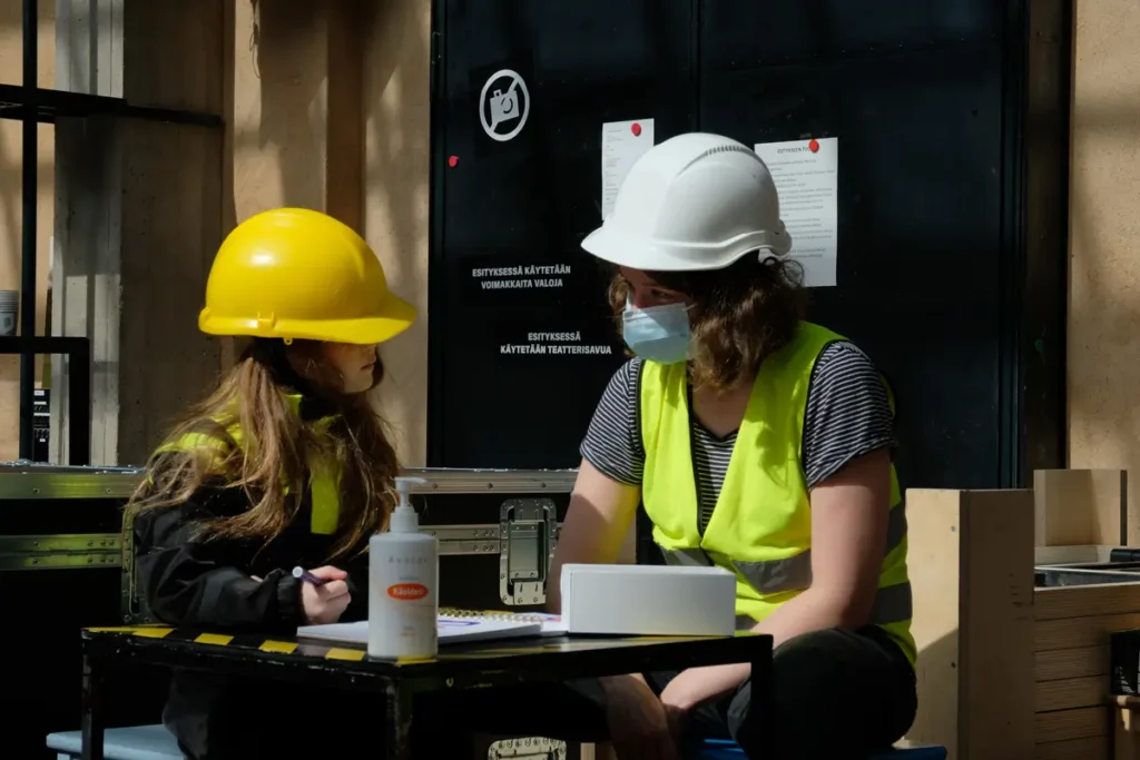 A photo showing a child and an adult sitting at a table wearing helmets, with a bottle of hand sanitiser on the table. The adult is wearing a face mask. The child has a pen and paper. They are looking at each other and appear to be having a conversation.