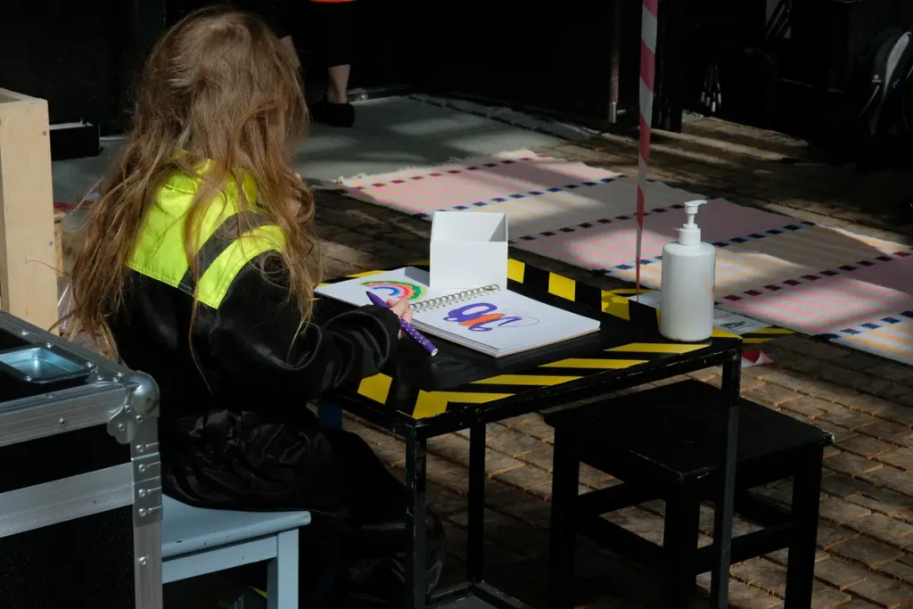 The pair of photos shows a child sitting at a black table, wearing a bright yellow reflective vest. In the photo on the left, the child is leaning over, drawing close to the drawing; in the photo on the right, the child is looking at the colourful drawing on the table. Other people and details of the space are visible in the background.