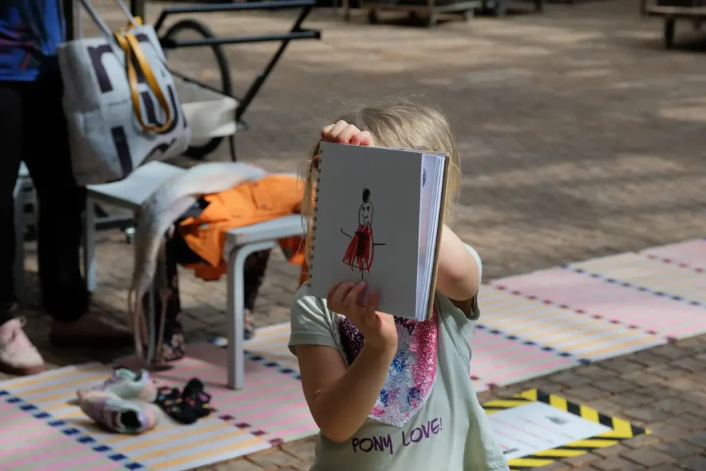 In the photo, a child is showing a drawing they made in the guestbook. The drawing depicts the researcher character wearing a red dress. The child is holding the drawing in front of their face so that their face is not visible in the photo. In the background, a chair, clothing, and colourful rugs on the outdoor floor are visible.