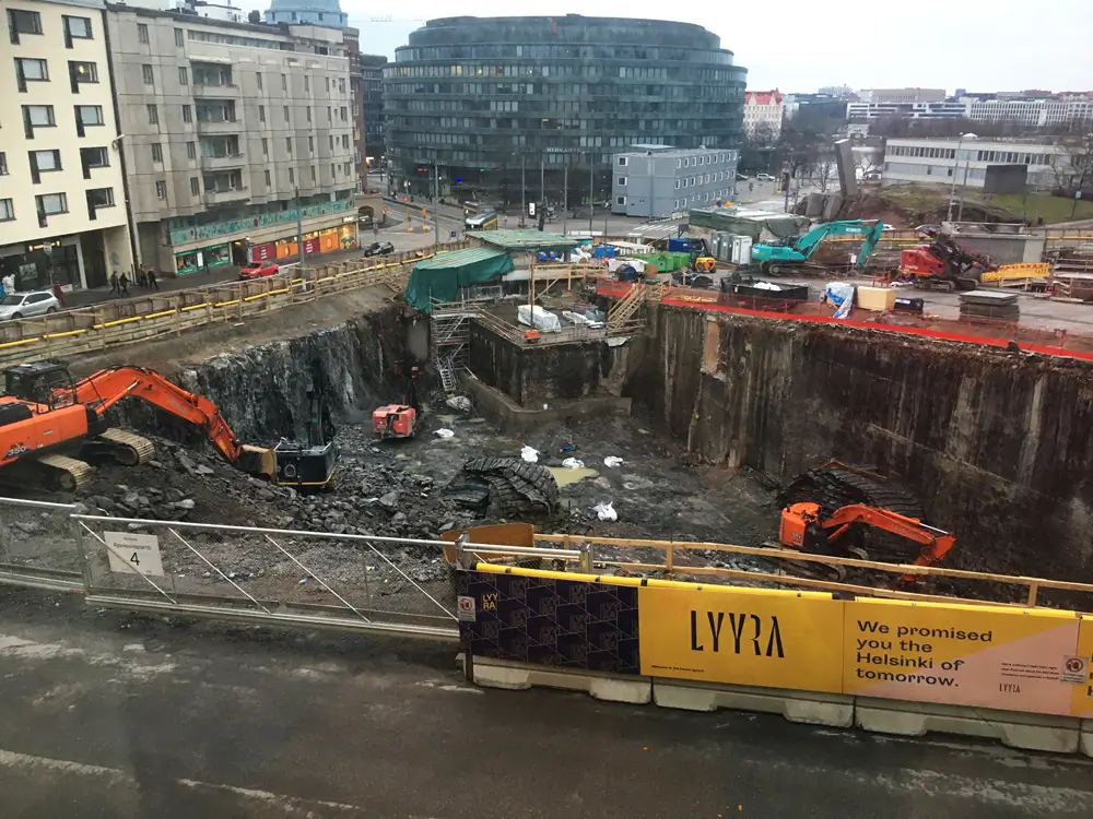 A photograph of a construction site in an urban setting, showing a large excavation and several excavators at work.