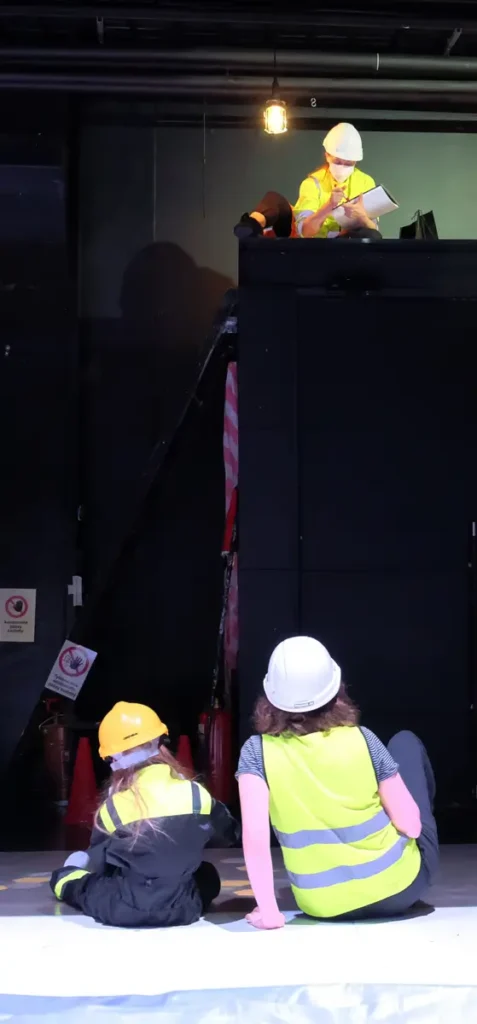 The photograph depicts a scene in which the researcher is sitting on a loft built above the doorway, while the participating child and adult are seated on the floor in front of it. The room features black walls, technical equipment, as well as caution tape and warning signs.
