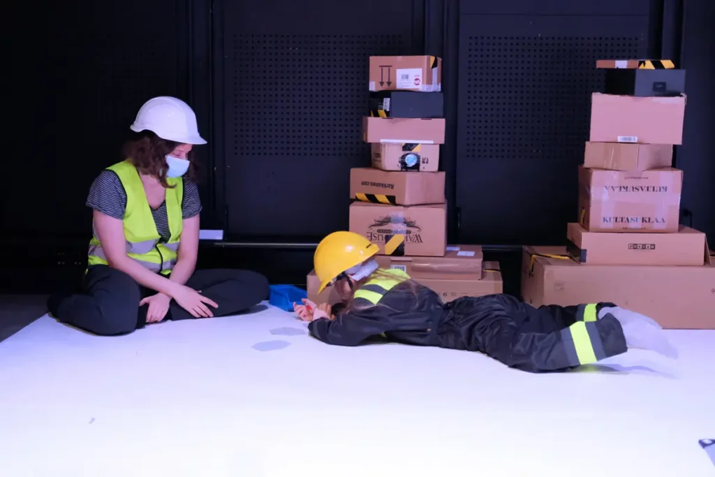 A photograph showing a child and an adult wearing helmets and construction-site safety gear. The child is lying on their stomach on the floor in front of a stack of cardboard boxes. The child is holding a pencil and has hexagonal pieces of paper in front of them. The adult is sitting on the floor next to the child, looking at the child.