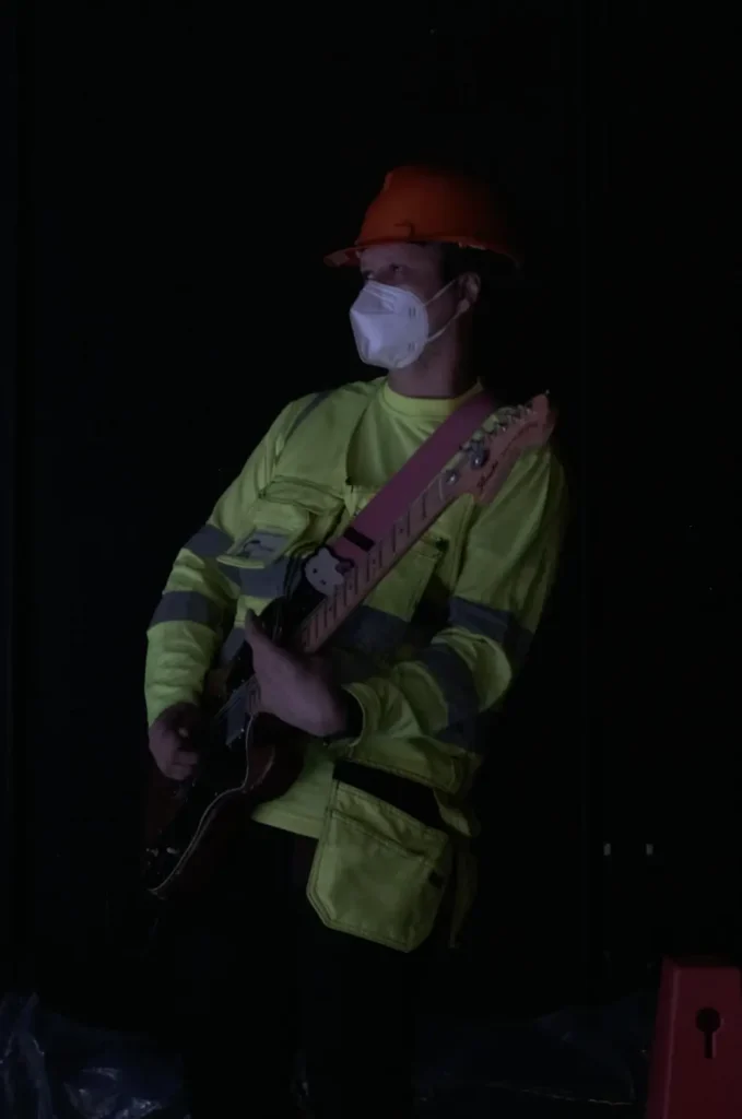 The photo shows a person wearing a yellow reflective vest, a red helmet, and a face mask, playing an electric guitar.