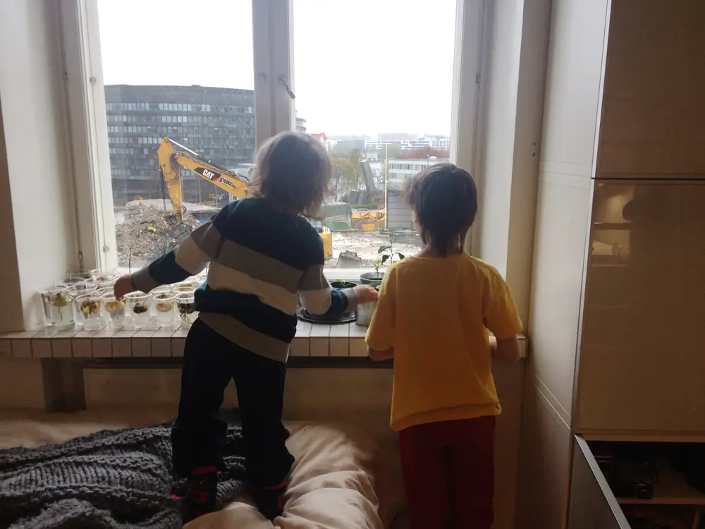 A photograph showing two children looking out the window at a construction site. An excavator and construction debris are visible in the background.