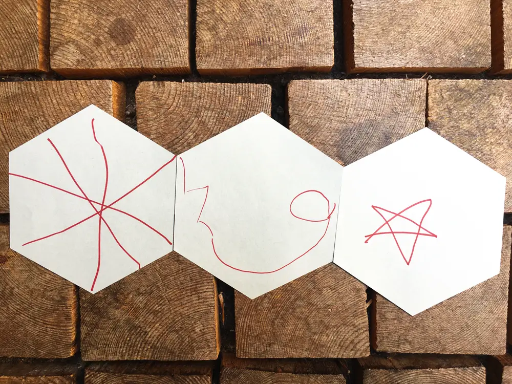 Three hexagonal pieces of paper with simple line drawings in red marker: radial lines on the left, a spiral-like line in the middle, and a star on the right.