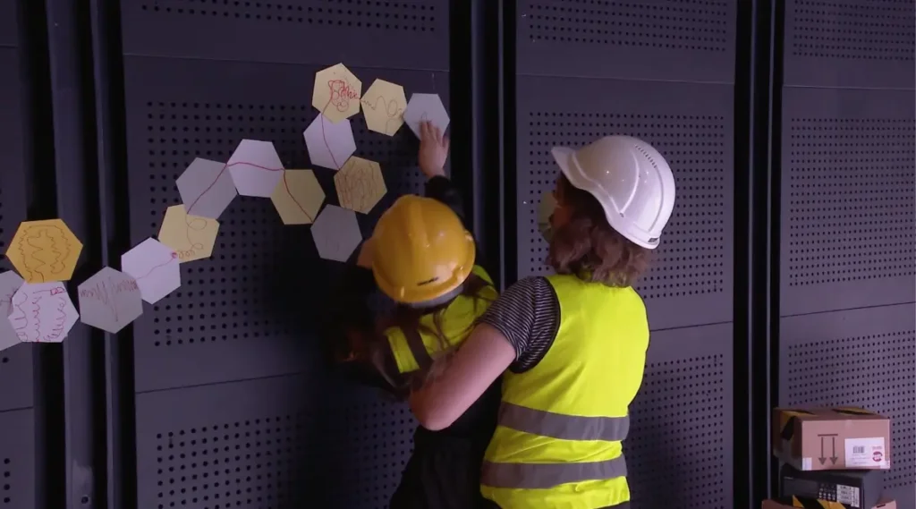 A photograph showing an adult and a child wearing safety helmets and yellow reflective vests. The adult is lifting the child, who is attaching paper hexagons to a black wall. The hexagons are connected to each other in a chain that forms a wave-like pattern.