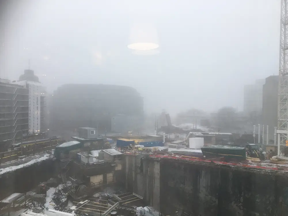 A photograph of a foggy construction site showing excavations, building materials, and cranes. Due to the fog, the buildings in the background are barely visible.