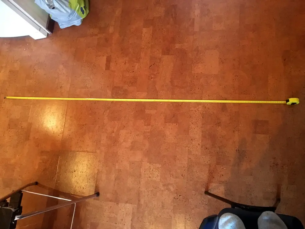 A photograph showing a long yellow measuring tape on the floor. Around the measuring tape, you can see the floor of the room, chairs, and part of the photographer&rsquo;s legs as they stand on a chair.
