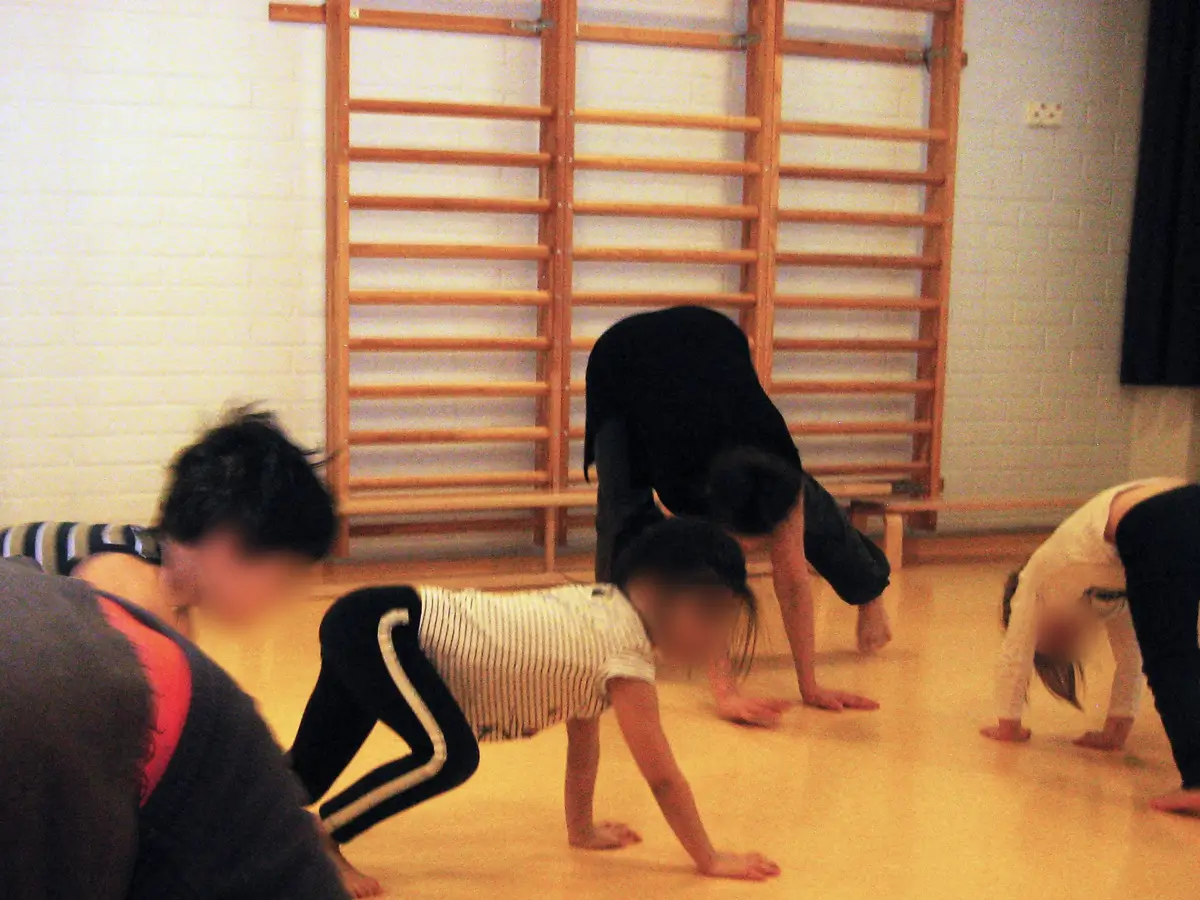 The photograph shows children and a researcher dancing in a gym, with wooden parallel bars visible in the background. Everyone is moving on the floor, using their arms and legs.